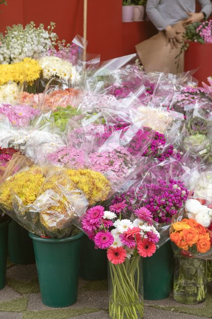 A vivid display of fresh flowers arranged in green buckets and clear plastic wrapping at a flower shop, featuring vibrant pink, white, yellow, and orange blooms such as gerbera daisies, chrysanthemums