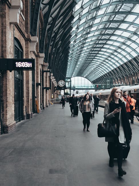This image depicts a busy train station platform with a high, arched glass ceiling allowing natural light to illuminate the space. The station features historic brick buildings with large windows and 