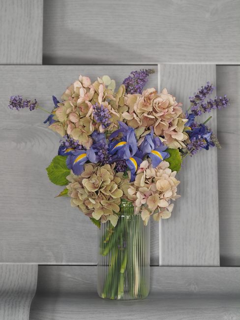 A floral arrangement featuring a bouquet of pastel pink and cream hydrangeas, complemented by vivid purple irises and small sprigs of lavender, arranged in a clear glass vase. The bouquet is placed on